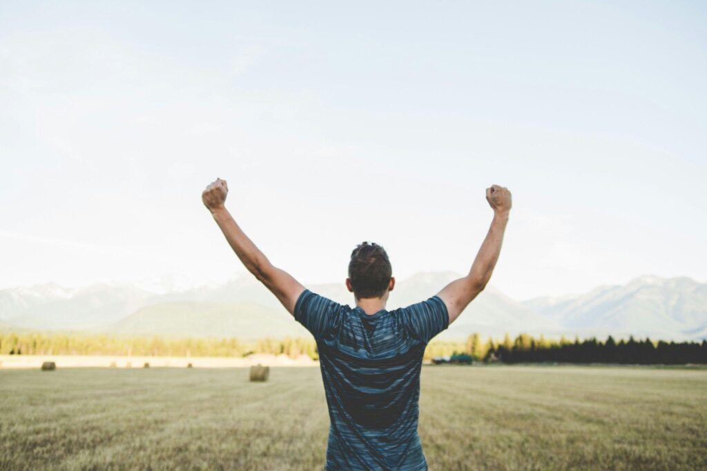 A man standing in an open field with mountains in the background, raising his arms in victory and strength, symbolizing personal transformation and resilience.